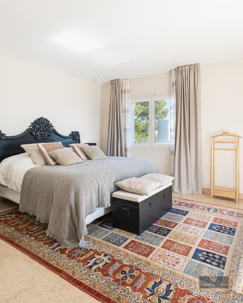 A bright master bedroom with an ornate black headboard, a colorful patterned rug, and travertine tile floors.