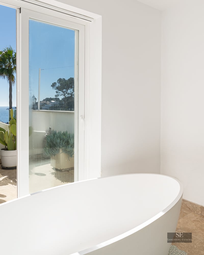 Modern bathroom featuring a white freestanding tub, Frida Kahlo portrait, and glass doors opening to a sea-view terrace.
