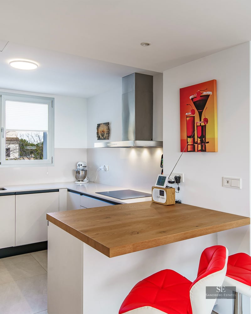 Bright modern kitchen featuring white cabinets, a wooden breakfast bar with red stools, and stainless steel appliances.
