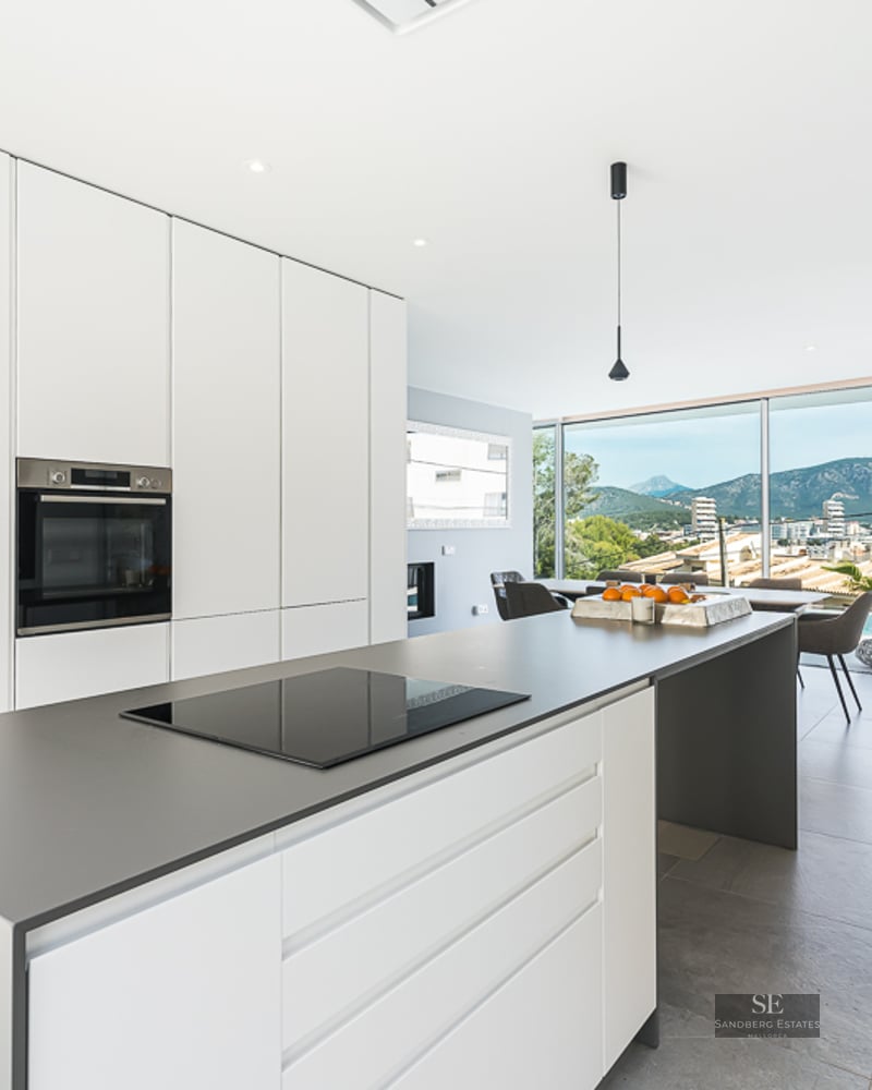 Bright white kitchen with a grey island, induction hob, and large windows overlooking mountains and a pool.