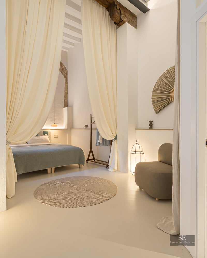 A serene master bedroom with flowing cream drapes, lantern lamps, and a glimpse into a stone-accented ensuite bathroom.