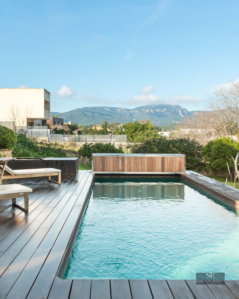 A rectangular swimming pool with a dark wood deck, white sun loungers, and a scenic mountain backdrop.