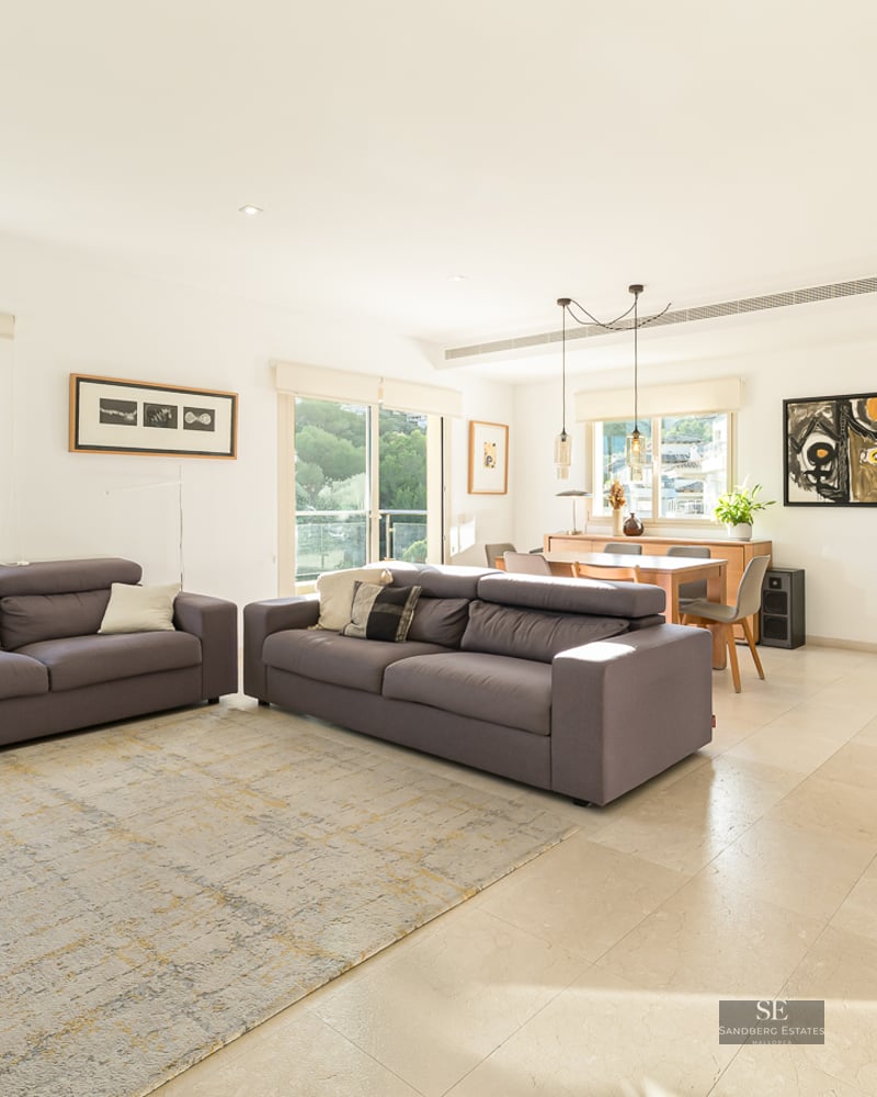 Spacious bright living room with two grey sofas, light tile flooring, and dining area in the background with large windows.