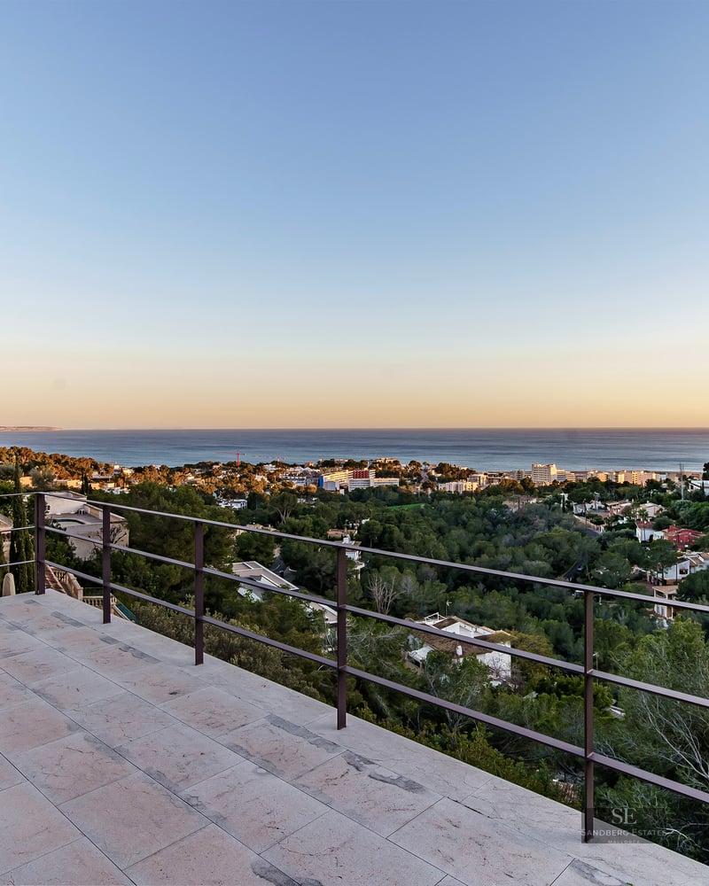 Wide stone terrace with dark metal railings overlooking a coastal town and the blue sea at sunset.