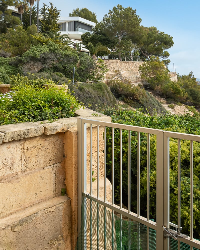 Stone steps and walls leading to a metal gate overlooking the Mediterranean sea and a distant island.