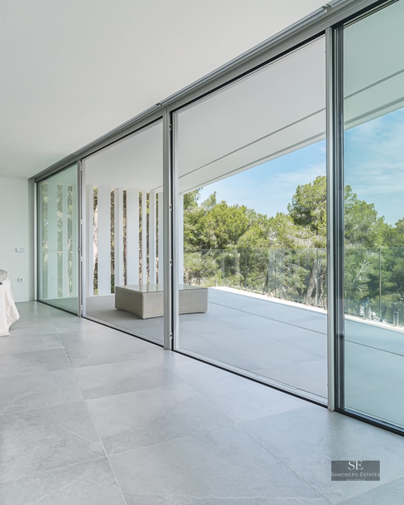 Bright minimalist bedroom with large sliding glass doors opening to a terrace with forest views.
