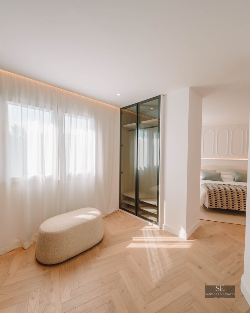 Bright dressing area with herringbone wood flooring, a beige ottoman, and glass wardrobe leading to a bedroom.