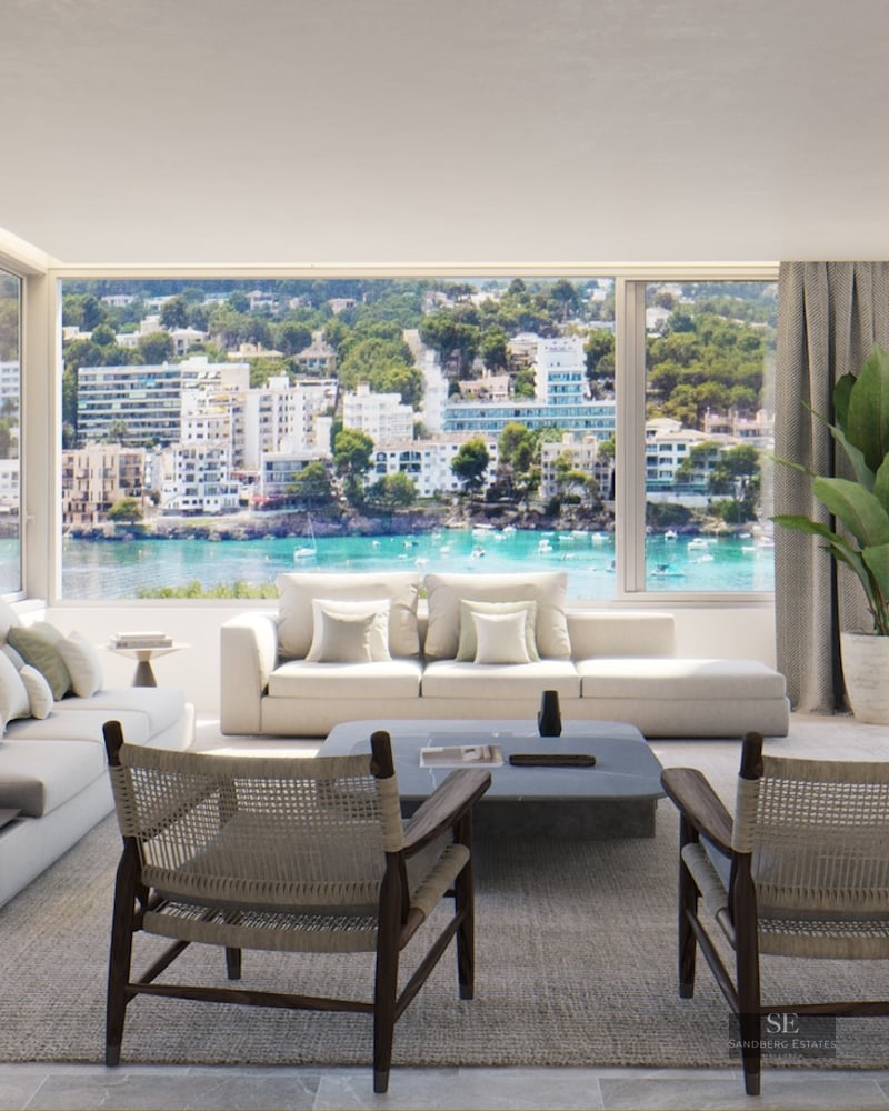 Bright living room with cream sofas and woven wood chairs facing a large window overlooking a Mediterranean bay.