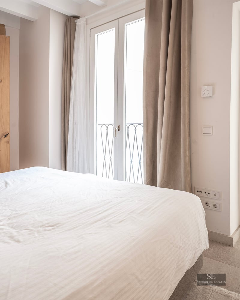 Chambre lumineuse avec lit blanc, grande armoire en bois et portes-fenêtres donnant sur un balcon.