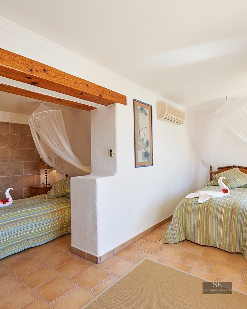 Bright bedroom featuring two twin beds with mosquito net canopies, exposed wooden beams, and terracotta tile floors.