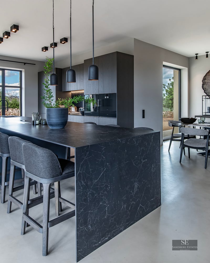 Modern open-plan kitchen with a dark marble island, grey stools, and a dining table under black designer pendant lights.