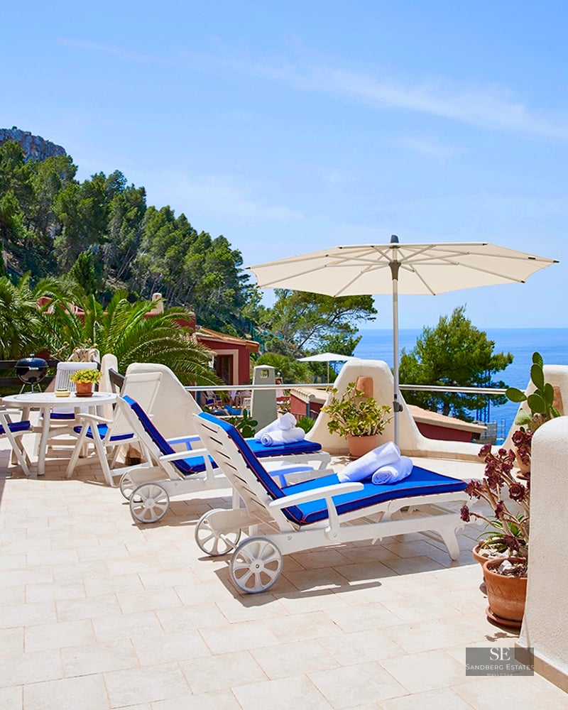 Sunny terrace with blue loungers, white umbrella, and panoramic views of the Mediterranean Sea and cliffs.