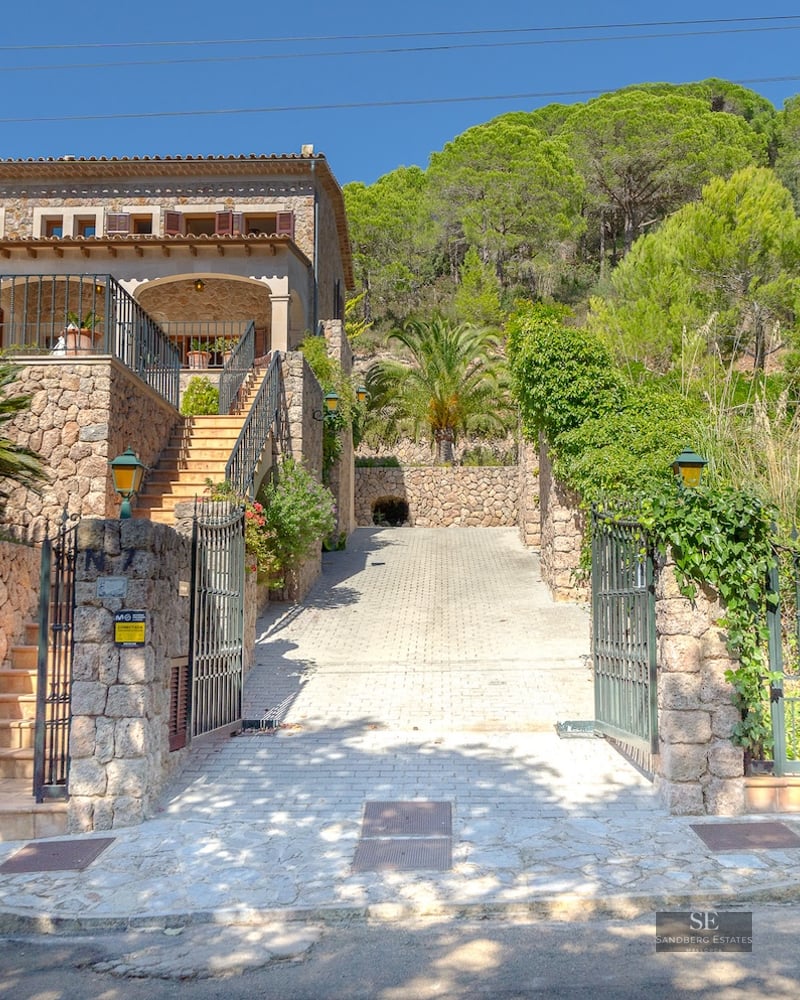 Two-story traditional stone house with iron gates, a paved driveway, and lush green trees against a bright blue sky.
