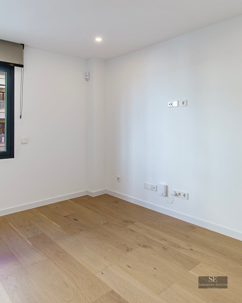 Pièce vide et lumineuse avec parquet en bois clair, murs blancs et une grande fenêtre avec store.