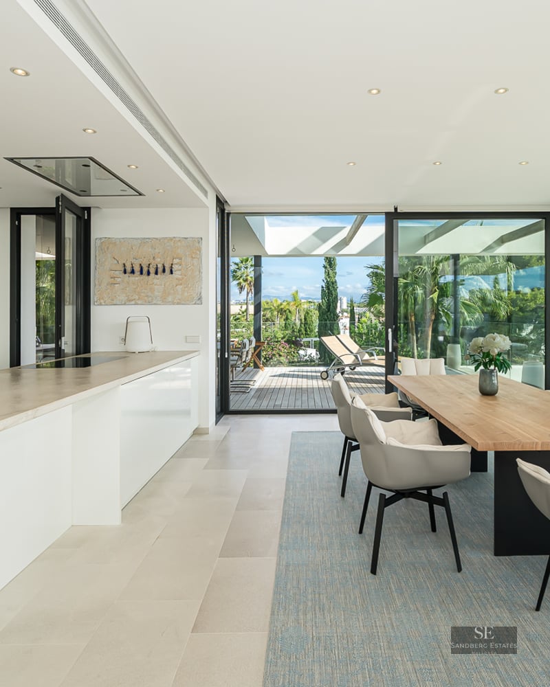 Modern dining room with a large wooden table, grey chairs, and a sleek white kitchen with floor-to-ceiling glass doors.