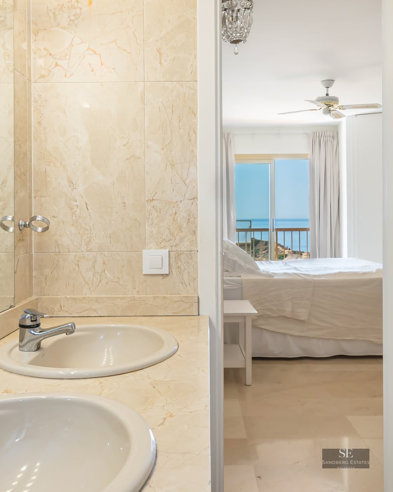 Master bathroom with beige marble double vanity looking into a bright bedroom with a sea view through an open door.