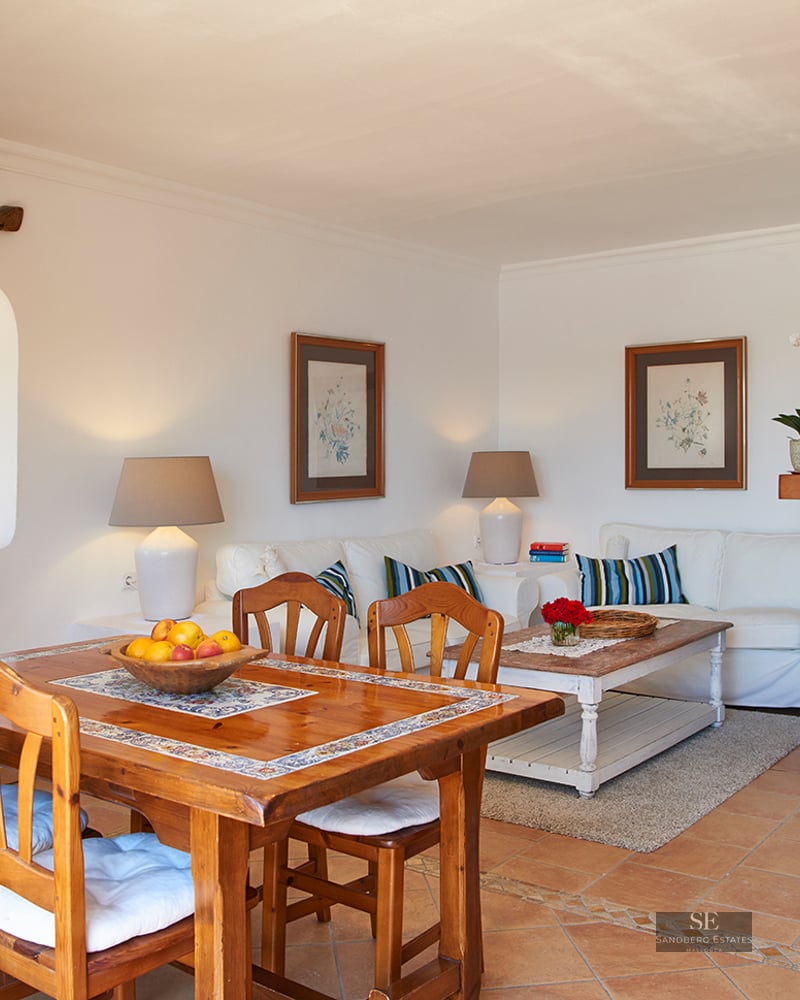 A bright living and dining space with terracotta tile floors, a wooden dining table, and a white sofa near a kitchen arch.