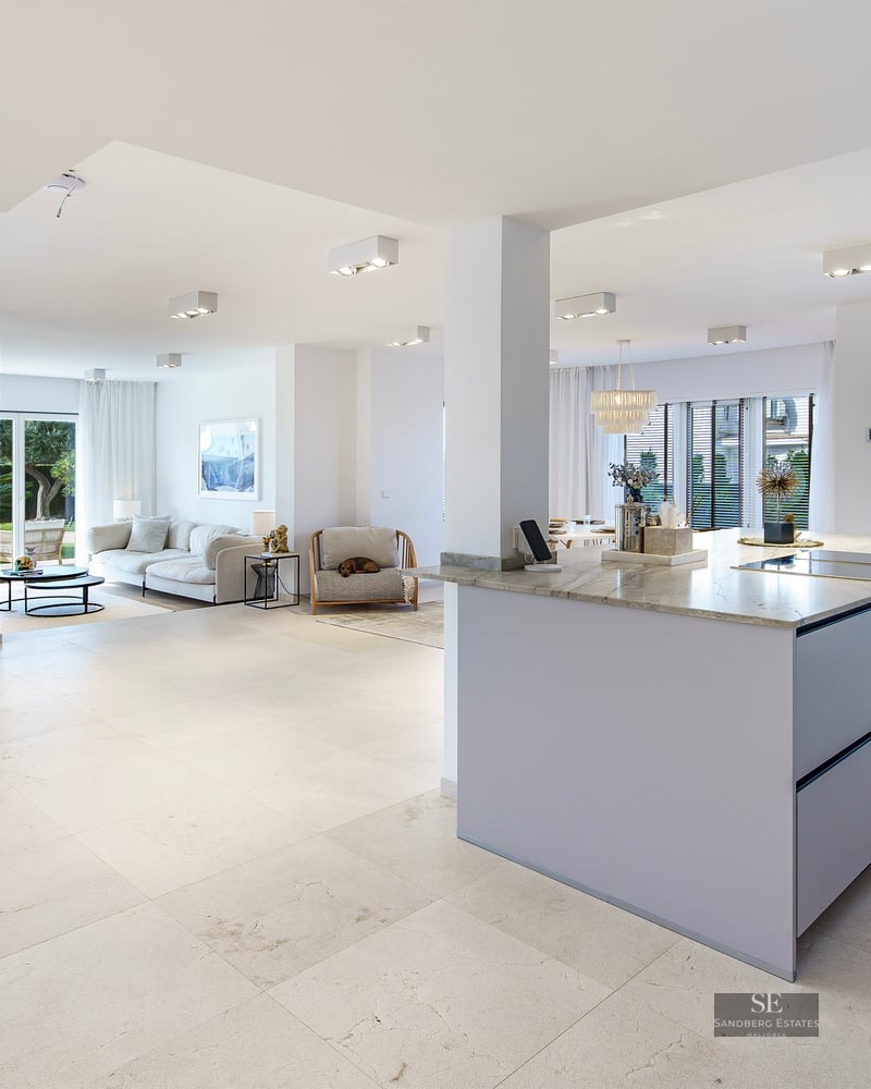 Bright open-plan living room with white sofa, marble kitchen island, and large glass doors leading to a garden.