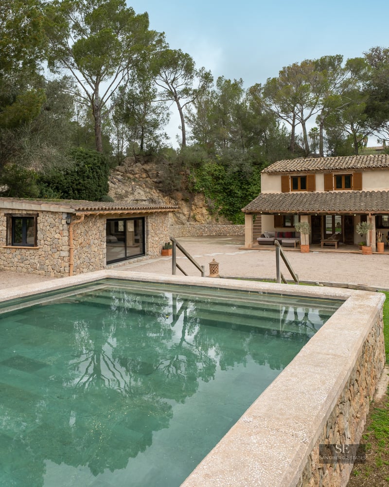Outdoor swimming pool with stone borders in front of a rustic Mediterranean villa surrounded by pine trees.