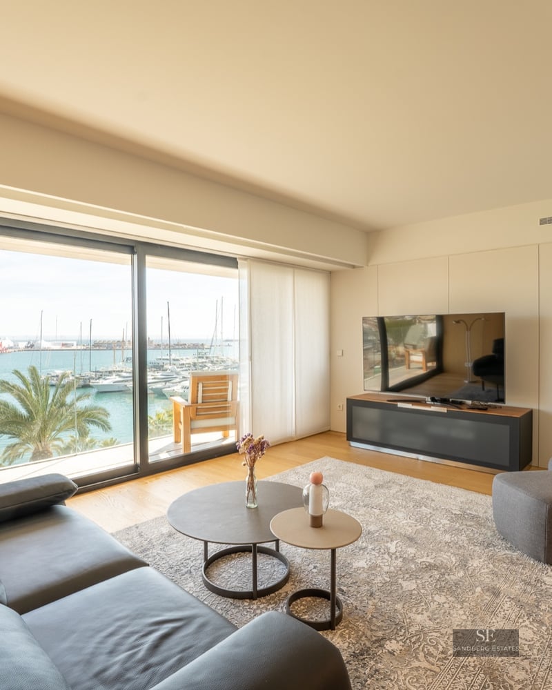 Bright modern living room with a black leather sofa and large windows overlooking a sunny marina with boats.