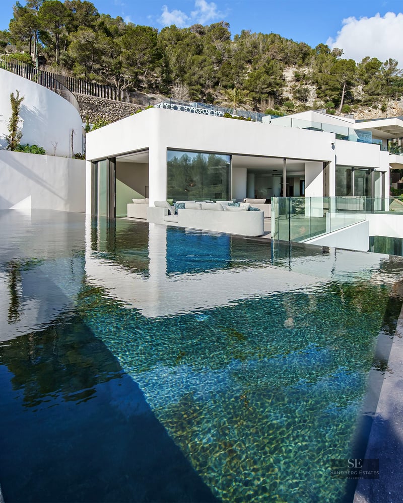 Modern infinity pool with clear blue water overlooking a white minimalist villa nestled in a green hillside.