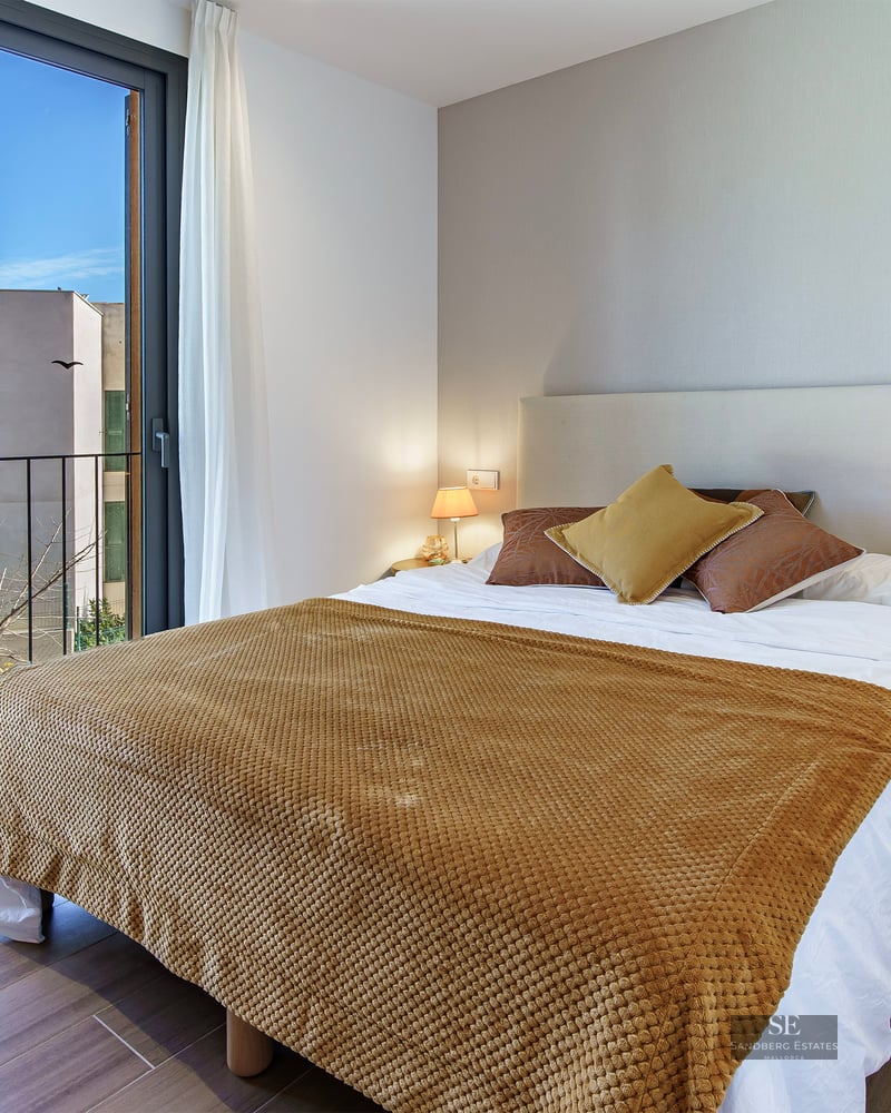 Bright bedroom featuring a double bed with an ochre yellow throw, a large glass sliding door, and a balcony view.