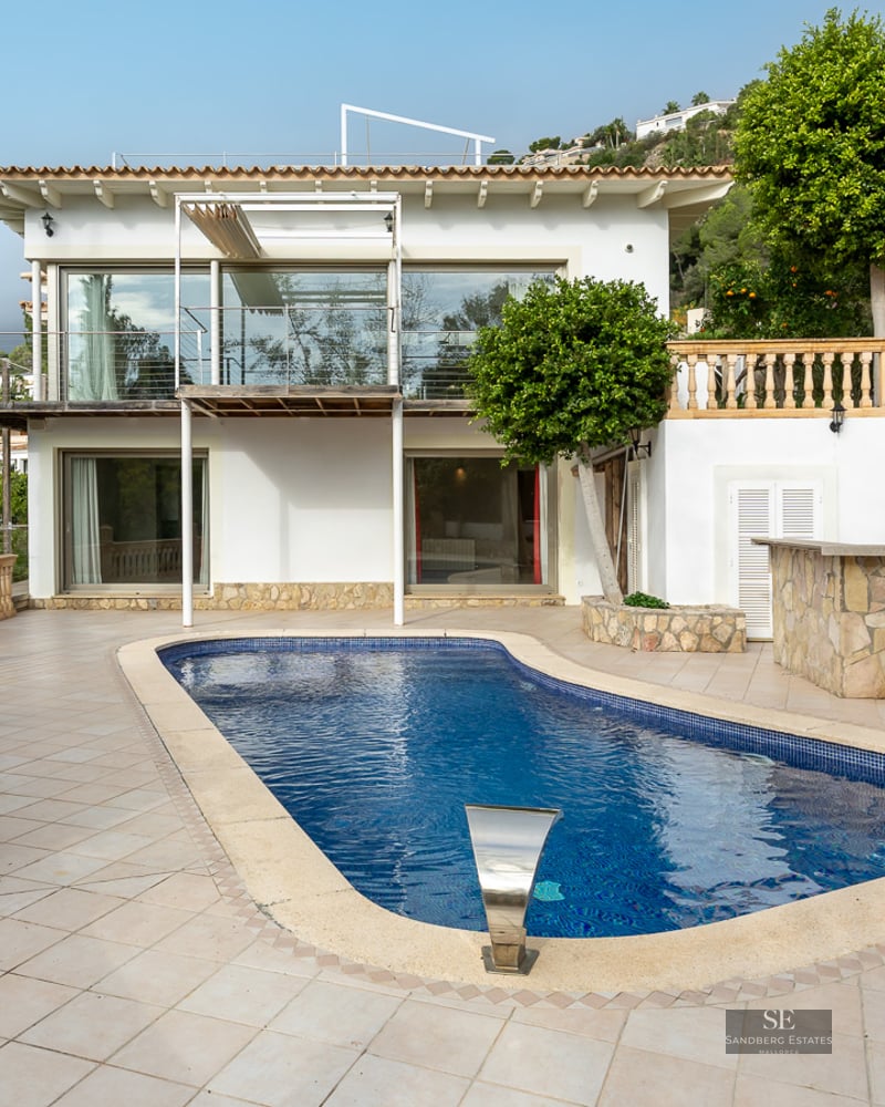 Blue swimming pool on a tiled terrace in front of a white Mediterranean villa with cypress trees.