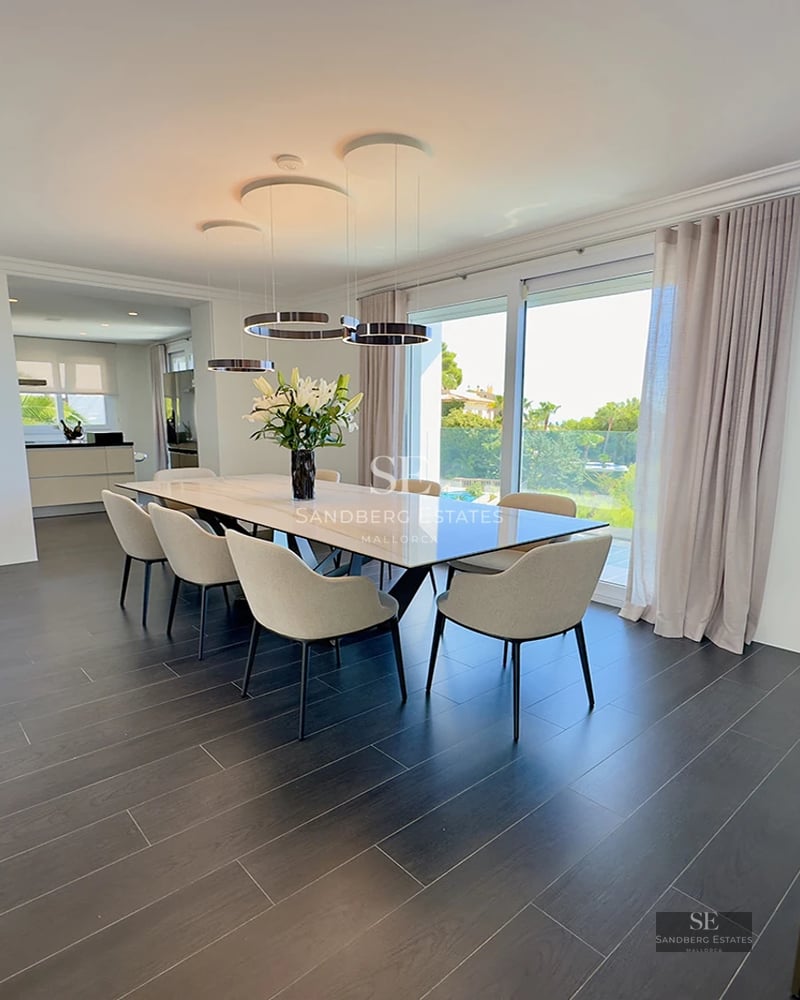 Modern dining room with a large marble table, cream chairs, dark floors, and floor-to-ceiling windows overlooking greenery.