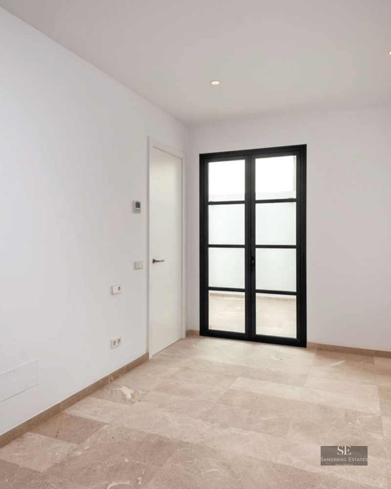 Empty room with white walls, beige stone floors, and black-framed glass doors leading outside.