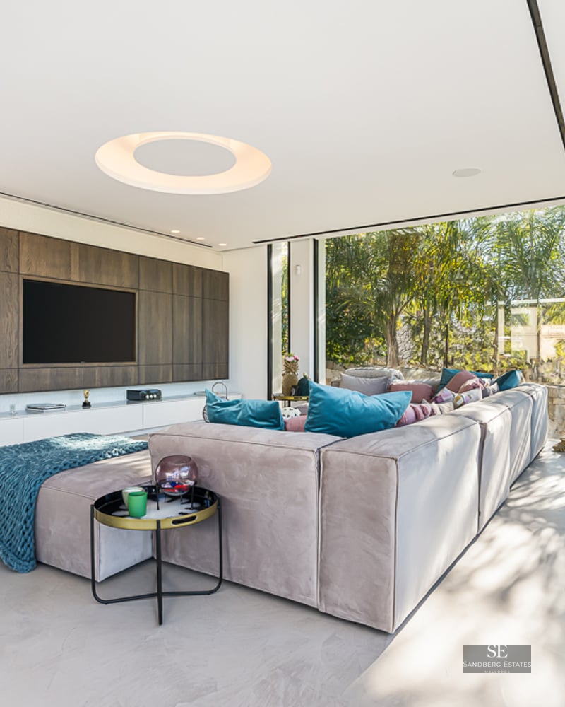 A bright contemporary living room with a large sectional sofa, wall-mounted TV, and floor-to-ceiling garden windows.