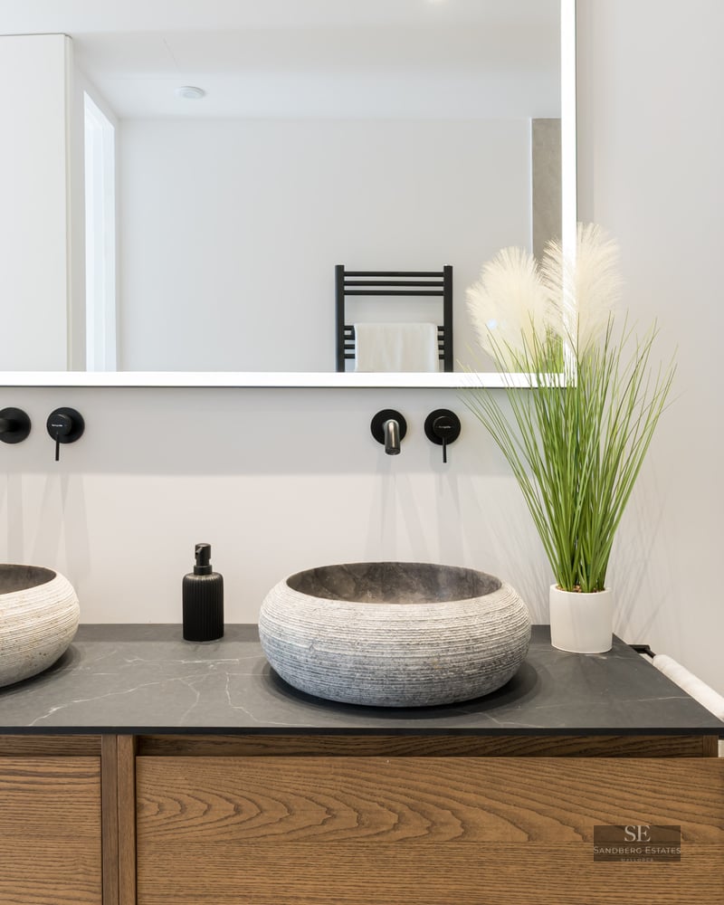 Modern bathroom vanity featuring two stone vessel sinks, black wall-mounted faucets, a backlit mirror, and wooden cabinets.