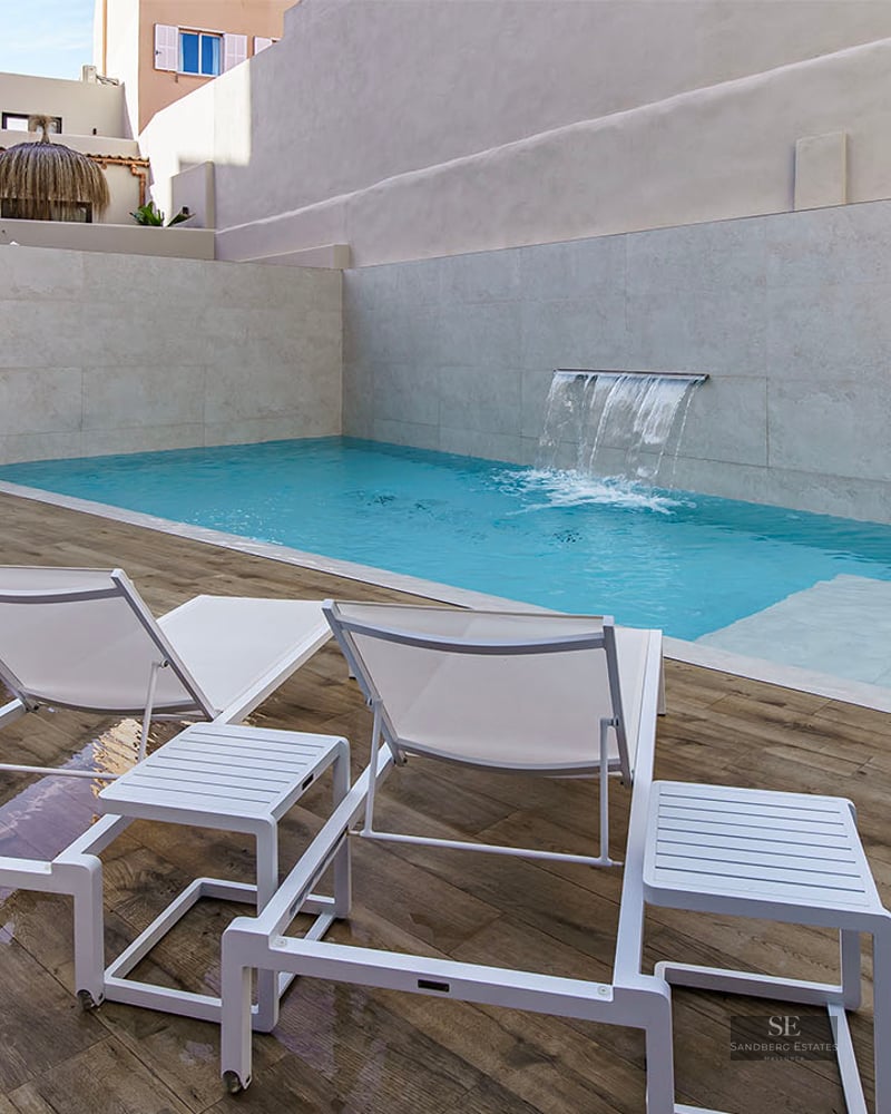Turquoise pool with integrated waterfall, white lounge chairs on wooden decking, and privacy stone walls.