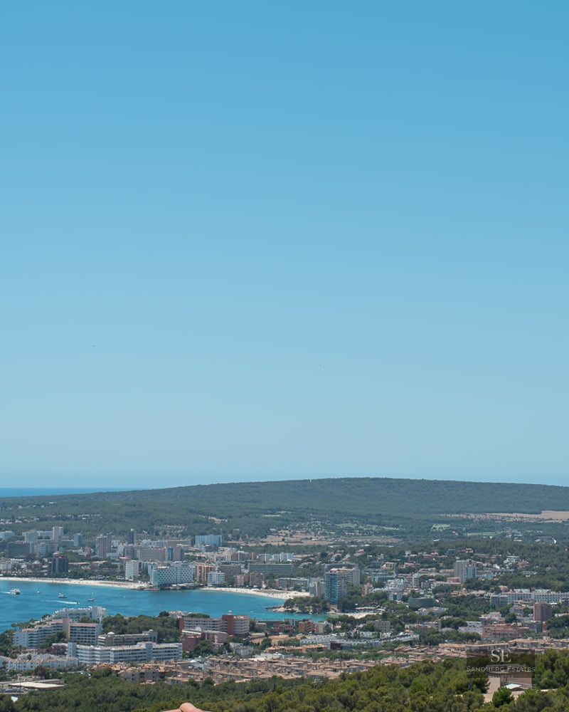 Panoramic elevated view of a blue Mediterranean bay, coastal town, and green hills under a clear blue sky.