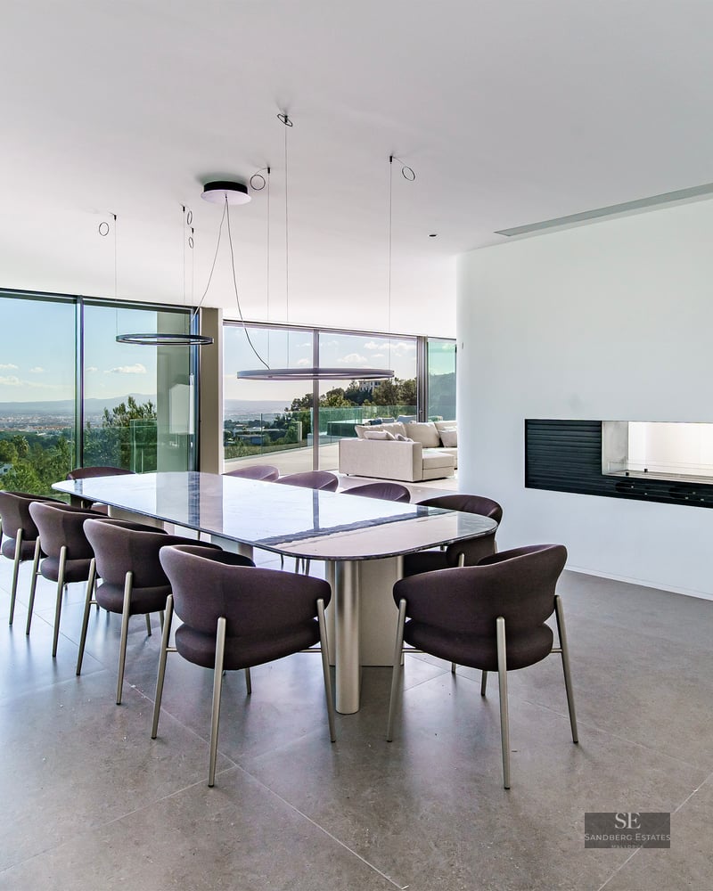 A modern dining room with a long table, dark grey chairs, and floor-to-ceiling windows overlooking a lush landscape.