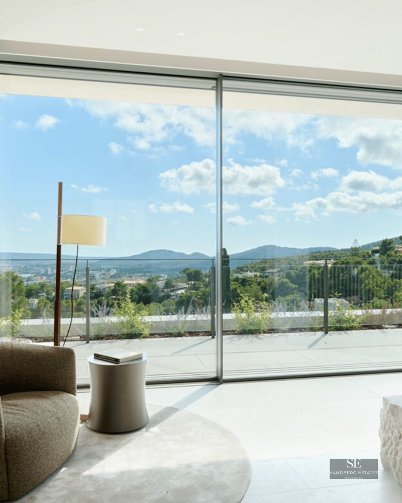 Modern interior featuring a textured armchair, stone fireplace base, and large windows overlooking green hills.