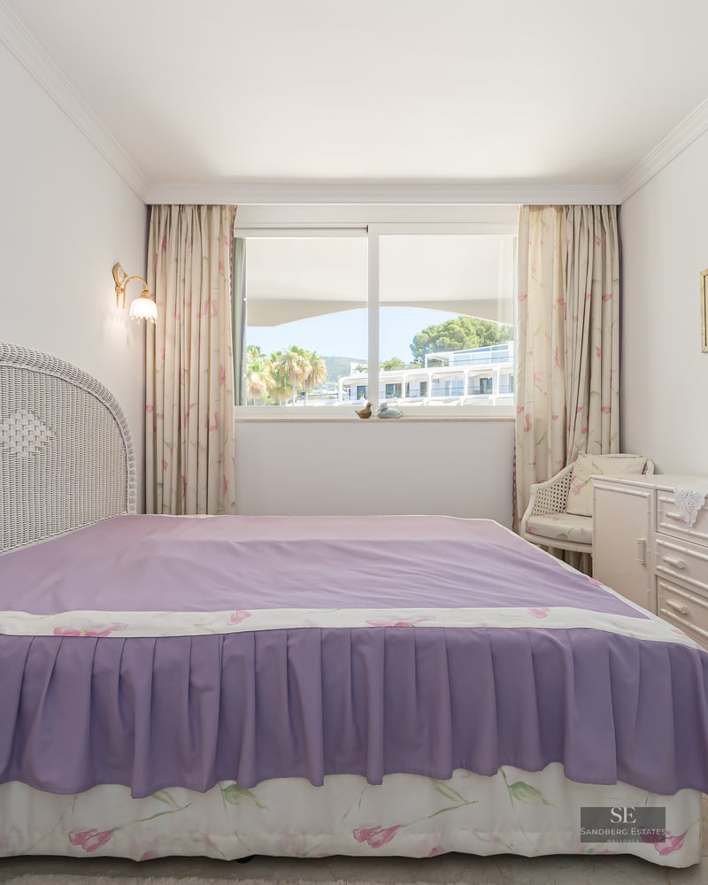 Bedroom featuring a white wicker headboard, purple bedspread, and a large window overlooking a sunny courtyard.