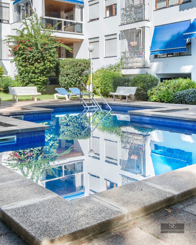 Rectangular blue pool surrounded by a stone terrace with loungers in front of a white apartment building.