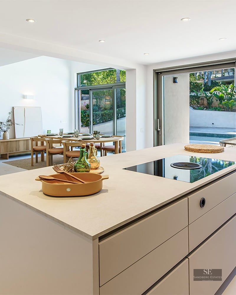 Bright, modern interior featuring a large kitchen island, dining area, sofas, and floor-to-ceiling windows leading to a pool.