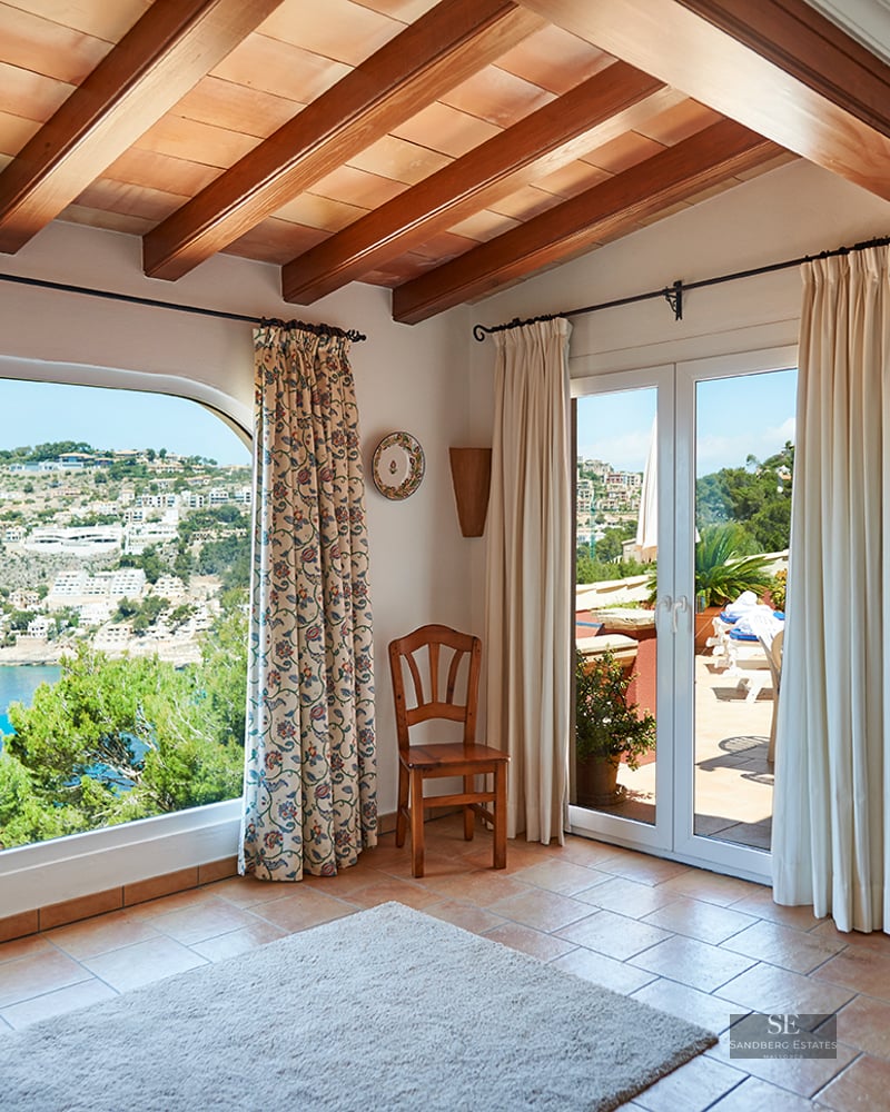 Interior room with exposed wood beams, terracotta floors, and a large arched window overlooking a coastal bay.