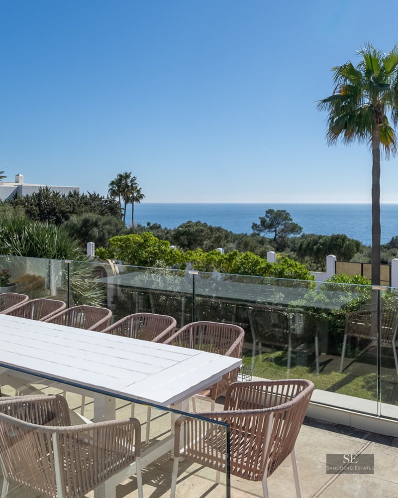A spacious outdoor terrace with a large white table, stylish chairs, and a glass railing overlooking the sea and palm trees.