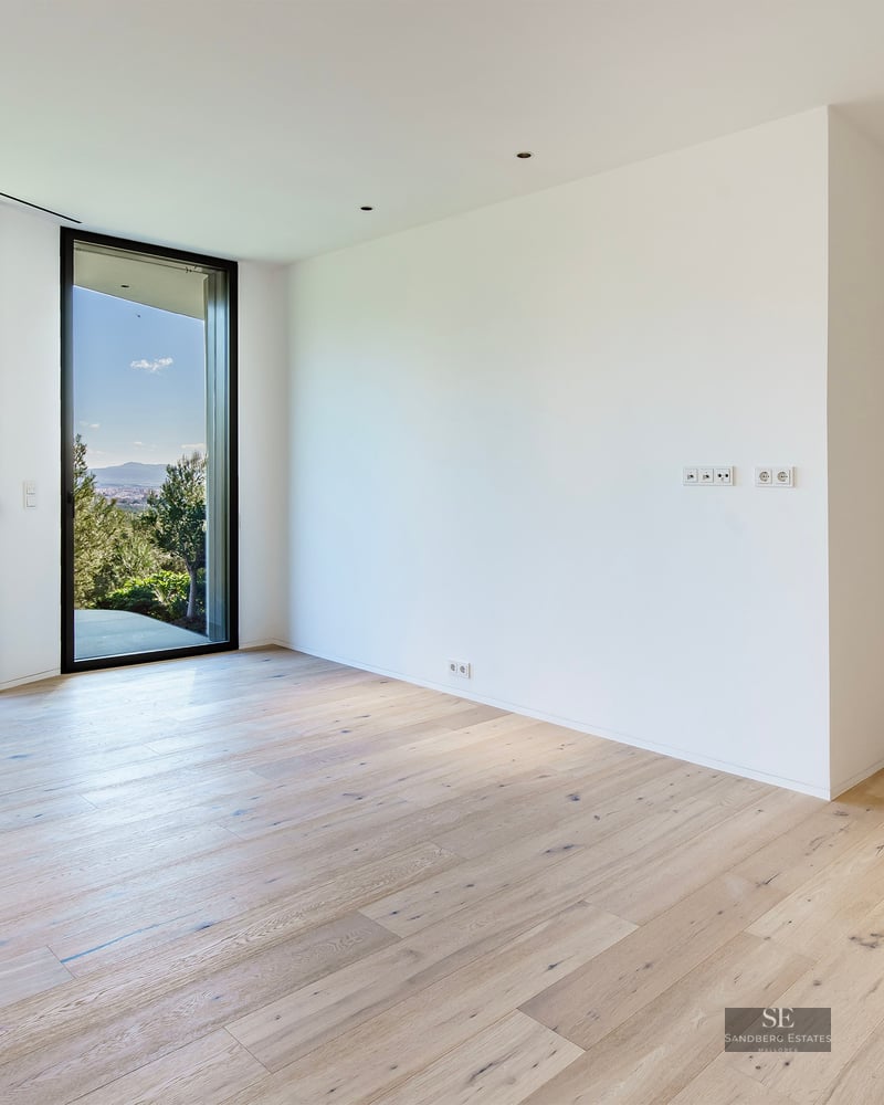 An empty room with light wood floors, white walls, and two large floor-to-ceiling windows looking out onto green trees.