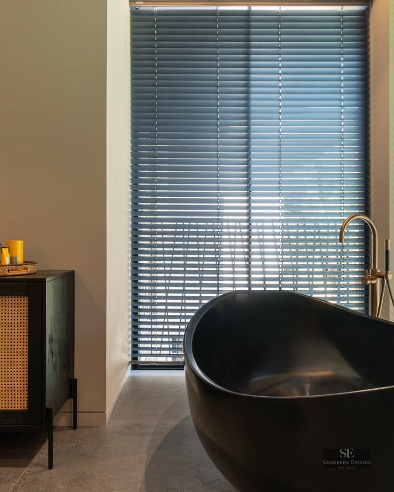 Luxury bathroom featuring a black oval bathtub, gold faucet, and a black rattan sideboard next to windows with blinds.