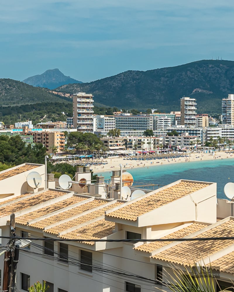 View overlooking a coastal bay with turquoise water, a sandy beach, white hotels, and green mountains in the background.