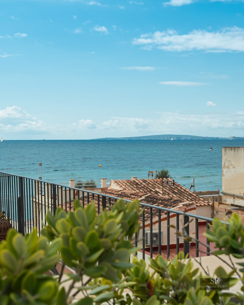 A view of the blue Mediterranean sea from a terrace with a black metal railing and terracotta roofs in the foreground.