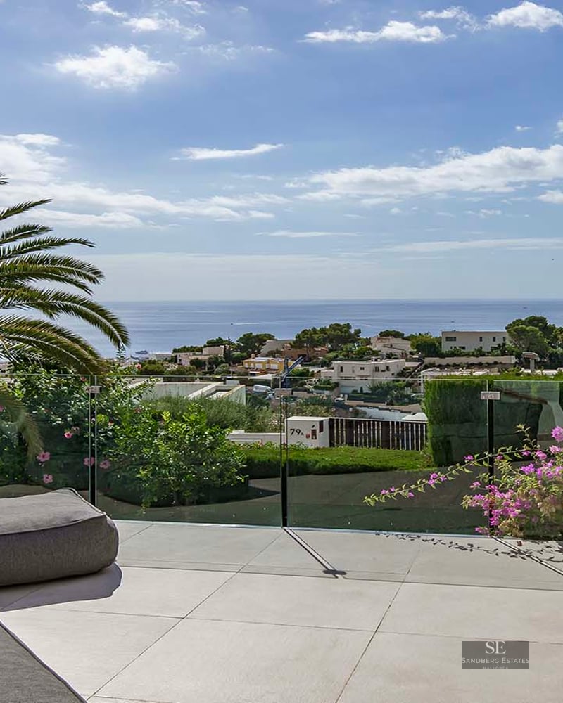 Luxury terrace with a grey outdoor sofa, glass railing, palm trees, and a stunning view of the sea and town.