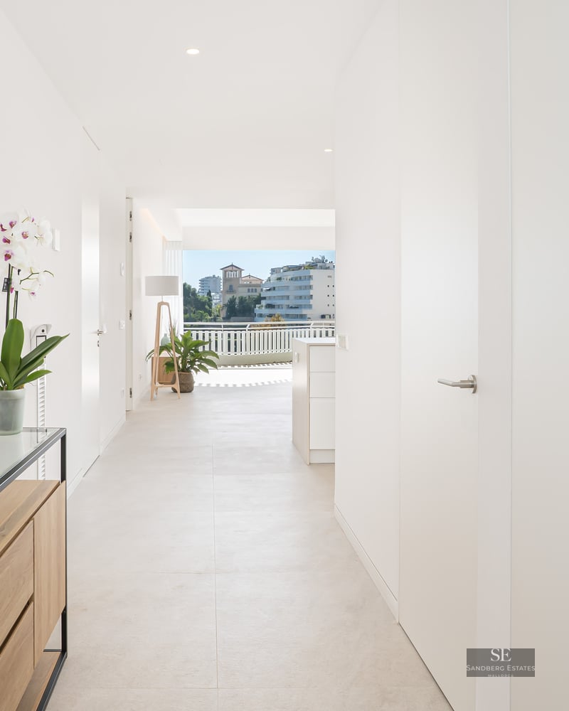 White hallway with gold sunburst mirror and wooden console, leading towards a sunny city view.