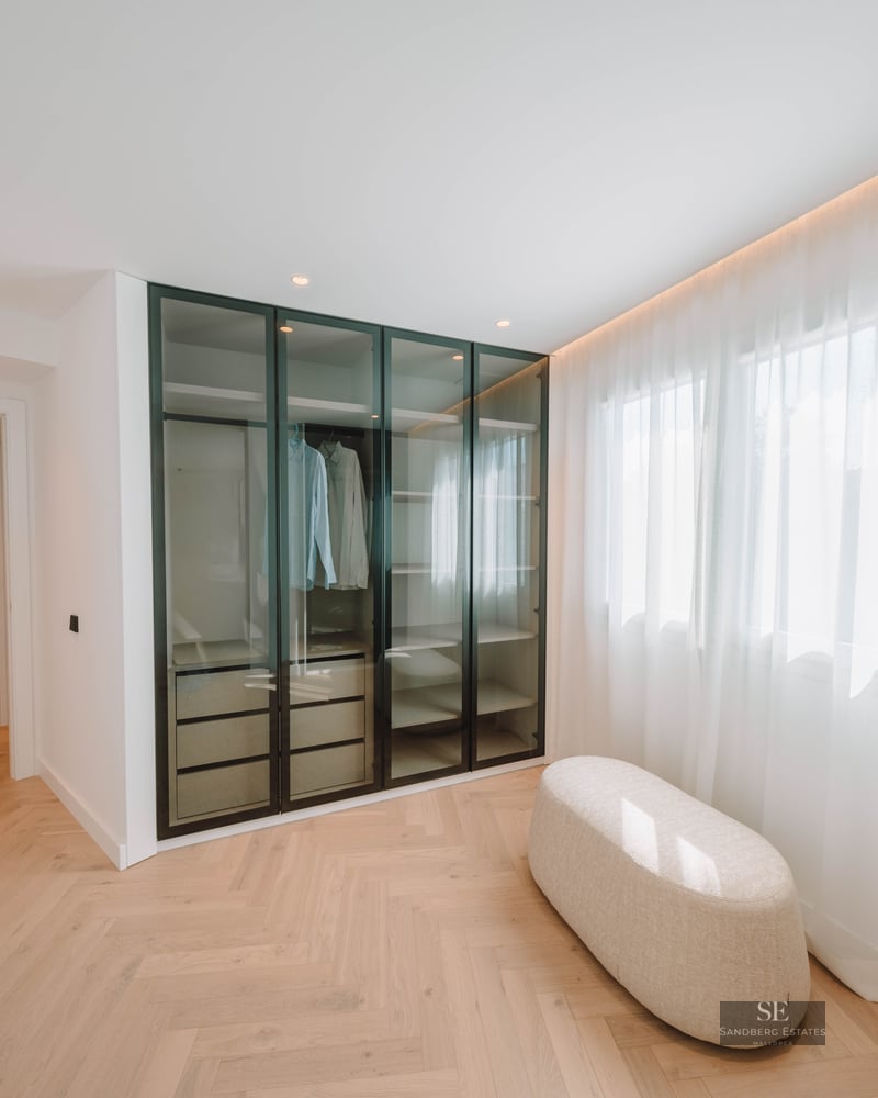 Modern walk-in closet with herringbone wood floors, glass wardrobe, and a central ottoman next to a window.