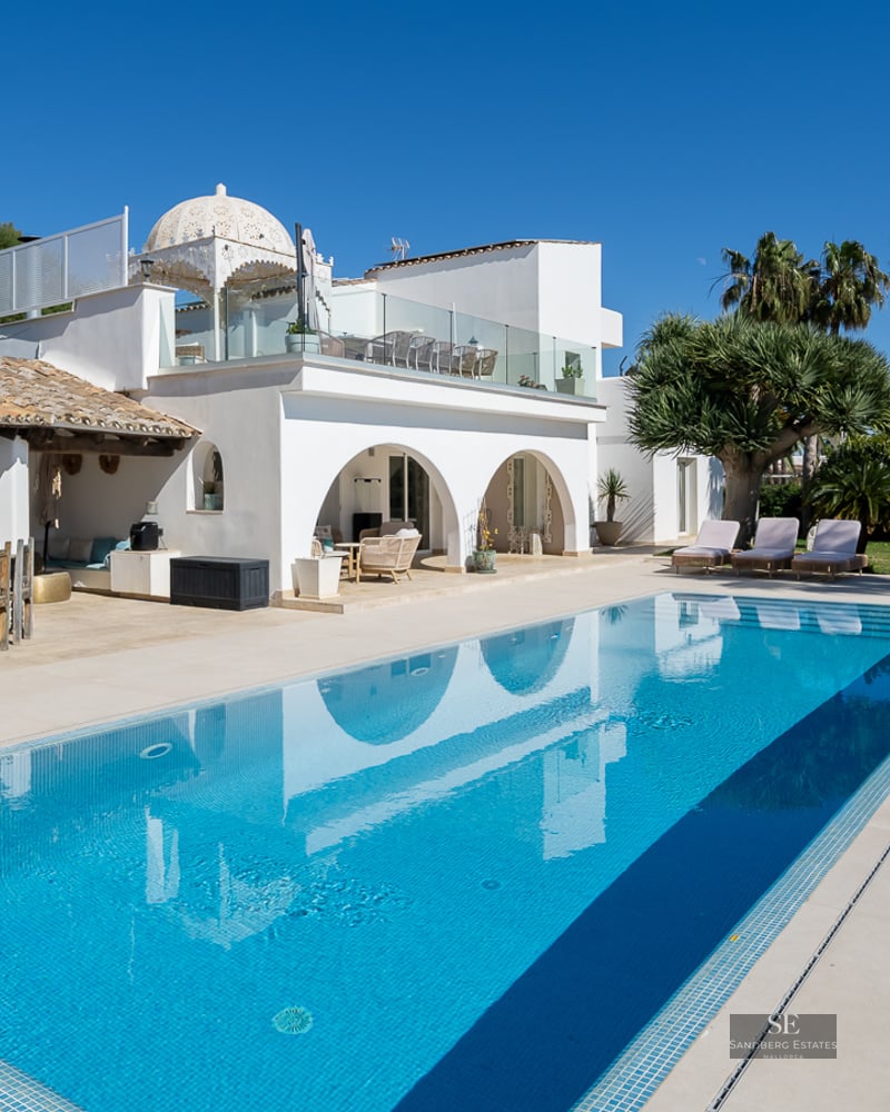A large blue swimming pool reflecting a white Mediterranean villa with arches, palm trees, and a Buddha statue.