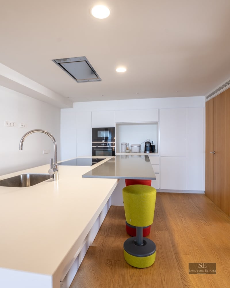 Bright modern kitchen interior featuring a large white island, sink, wooden floors, and built-in appliances.