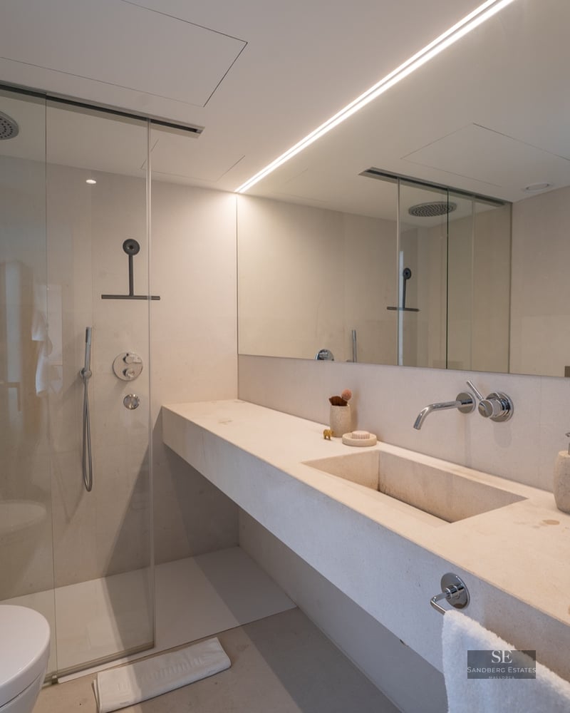 Modern bathroom with cream stone vanity, glass walk-in shower, large mirror, and linear lighting.